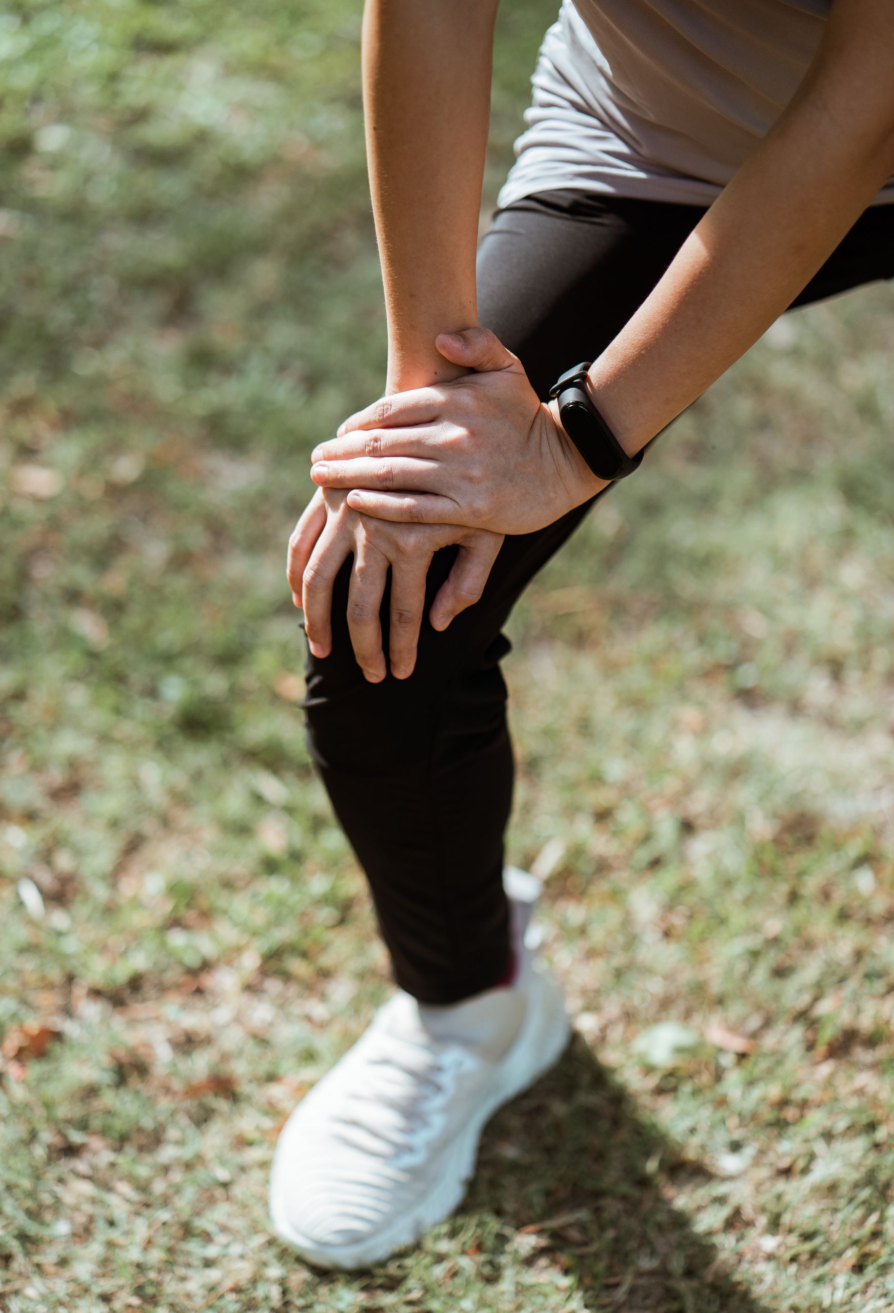 Crop anonymous slim sportswoman in black leggings and white sneakers stretching legs before jogging while warming up on green meadow in sunlight